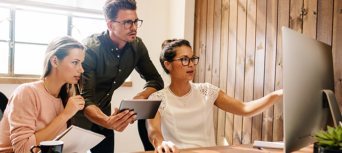 Three people look at something on a computer monitor.